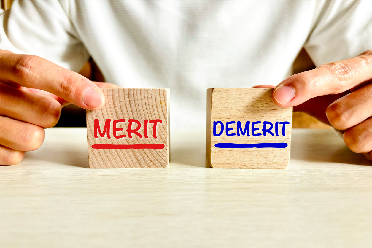 机の上に置かれた「MERIT（メリット）」と「DEMERIT（デメリット）」と書かれた木のブロックを、それぞれ左右の手で掴んでいる画像